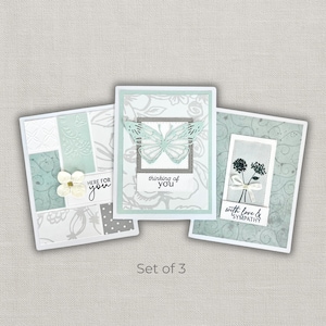 May include: Set of three handmade greeting cards. Each card features a unique design with floral and butterfly accents in shades of white, gray, and teal. Text includes "Here for you", "thinking of you", and "with love & sympathy".