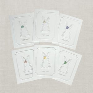 May include: A collection of white Easter greeting cards. Each card features a cut-out bunny design with a small flower accent in various colors. The words "happy easter" are printed at the bottom of each card.