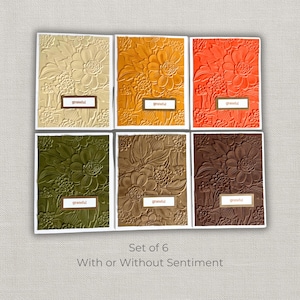 May include: A set of six embossed floral greeting cards in various colors: cream, orange, red, green, brown, and dark brown. Each card has a rectangular label that reads "grateful". The cards are displayed on a light gray surface.