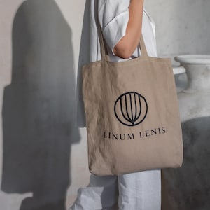 May include: A beige tote bag with a black circular logo and the text "LINUM LENIS". The bag is held by a person. The design is simple, made of natural fabric. The background includes a large vase and a shadow.