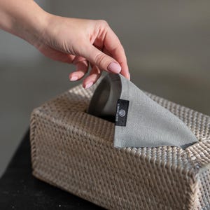 Könnte beinhalten: Ein grauer Leinen-Tischtuchbezug mit dem Text "LINUM LENS" in Schwarz. Der Bezug befindet sich auf einer geflochtenen Weiden-Tischtuchbox.