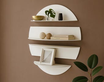 May include: A set of four white, modern wall shelves in a unique, curved design. The shelves are arranged in a semi-circle, with decorative items like a gold bowl, a black vase, and a framed picture. A green plant is visible on the right.
