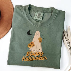 Puede incluir: Camiseta verde oliva con un diseño de fantasma con girasoles, una luna creciente y la frase "Happy Halloween" en naranja. Fabricada en material Comfort Colors.