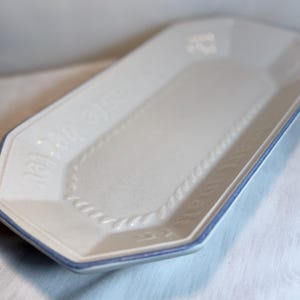 May include: An off-white ceramic serving platter with a blue trim. The rectangular platter has an octagonal shape and features embossed text and a rope-like border design. The words "Isle de Butler" are visible.
