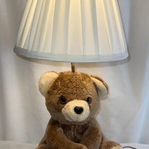 Könnte beinhalten: Teddybär-Lampe mit einem braunen Plüschbären mit cremefarbenen Akzenten, gekrönt von einem hellblauen und weißen Lampenschirm. Die Lampe steht auf einem Holzsockel. Ideal für ein Kinderzimmer, ist die Lampe eine dekorative Leuchte.