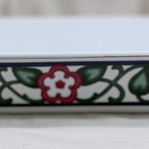 Puede incluir: Fuente rectangular blanca para hornear con un borde floral decorativo. El borde presenta una flor roja, hojas verdes y un diseño azul y blanco. El plato tiene un borde ligeramente festoneado.