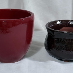 May include: Two ceramic planters. One is a glossy, deep red, rounded shape. The other is a smaller, dark brown, textured pot with a slightly irregular shape. Both are empty and ready for plants.