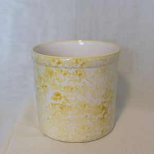 Puede incluir: Un recipiente cilíndrico de cerámica con un patrón moteado amarillo y blanco. El borde es amarillo liso y el interior es blanco. Este artículo de cocina podría usarse como un soporte para utensilios o un jarrón decorativo.