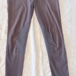 May include: Gray riding pants with reinforced knee patches and stirrup straps. The pants have a button and zipper closure, belt loops, and the brand name "Devon Aire" printed on the waistband. The pants are made of a durable material.