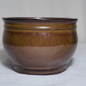 Puede incluir: Una maceta de cerámica hecha a mano, de forma redondeada y con una amplia abertura. La maceta presenta un degradado de colores, pasando de un borde marrón oscuro a una base marrón más clara. La maceta es adecuada para plantas o uso decorativo.
