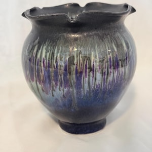 May include: A ceramic vase with a ruffled rim and a bulbous body. The vase has a dark, iridescent glaze with streaks of purple, blue, and green. The base is a darker shade of blue.