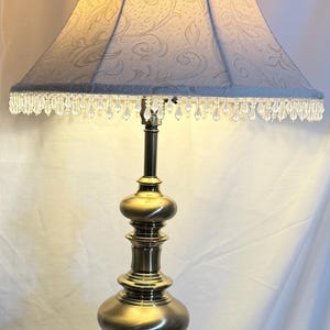 Puede incluir: Lámpara de mesa de estilo vintage con base de latón y pantalla azul claro. La pantalla tiene un estampado floral y está adornada con cuentas de cristal colgantes. La lámpara está encendida, iluminando la pantalla.