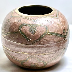 May include: A round, decorative container with a metallic pink finish and gold trim. The container features a heart and floral design in green and gold. The top opening is black.