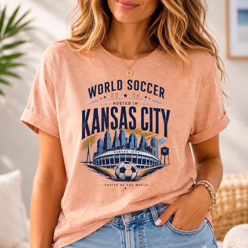 May include: A peach-colored short-sleeve t-shirt featuring a dark blue graphic print that reads World Soccer 2026 Hosted In Kansas City. The design includes a city skyline, a stadium, a soccer ball, and the text United By The World.