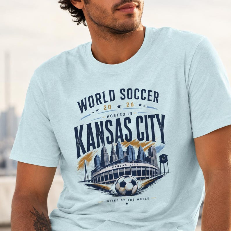 May include: A light blue t-shirt featuring a graphic with the text "WORLD SOCCER 20 26 HOSTED IN KANSAS CITY" and "UNITED BY THE WORLD". The design includes a stylized illustration of the Kansas City skyline, a stadium, and a soccer ball.
