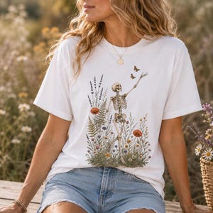 May include: A white short-sleeve t-shirt featuring a graphic print of a skeleton standing amidst a field of colourful wildflowers, ferns, and lavender, with two small brown butterflies flying above the skeleton's raised hand.