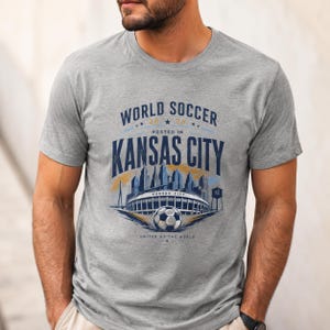 May include: A heather gray short-sleeve t-shirt featuring a graphic print that reads WORLD SOCCER 2026 HOSTED IN KANSAS CITY. The design includes a blue skyline illustration, a stadium, a soccer ball, and the text UNITED BY THE WORLD.