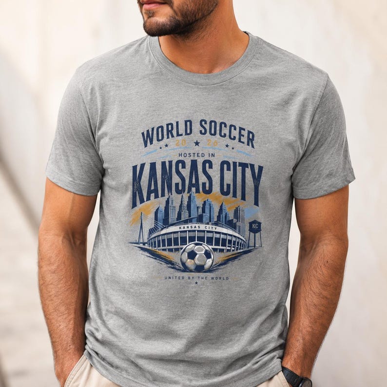 May include: A heather gray short-sleeve t-shirt featuring a graphic print that reads WORLD SOCCER 2026 HOSTED IN KANSAS CITY. The design includes a blue skyline illustration, a stadium, a soccer ball, and the text UNITED BY THE WORLD.