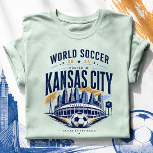 May include: A light green t-shirt featuring a graphic with the text WORLD SOCCER 2026 HOSTED IN KANSAS CITY UNITED BY THE WORLD. The design includes a soccer ball, a stadium, a city skyline, and a water tower labeled KC in dark blue and gold.