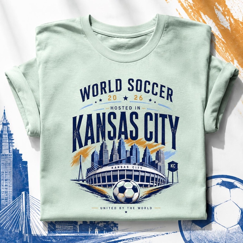 May include: A light green t-shirt featuring a graphic with the text WORLD SOCCER 2026 HOSTED IN KANSAS CITY UNITED BY THE WORLD. The design includes a soccer ball, a stadium, a city skyline, and a water tower labeled KC in dark blue and gold.