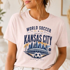 May include: A light pink t-shirt featuring a graphic with the text WORLD SOCCER 2026 HOSTED IN KANSAS CITY UNITED BY THE WORLD. The design includes a blue skyline illustration, a stadium, a soccer ball, and a water tower labeled KC.