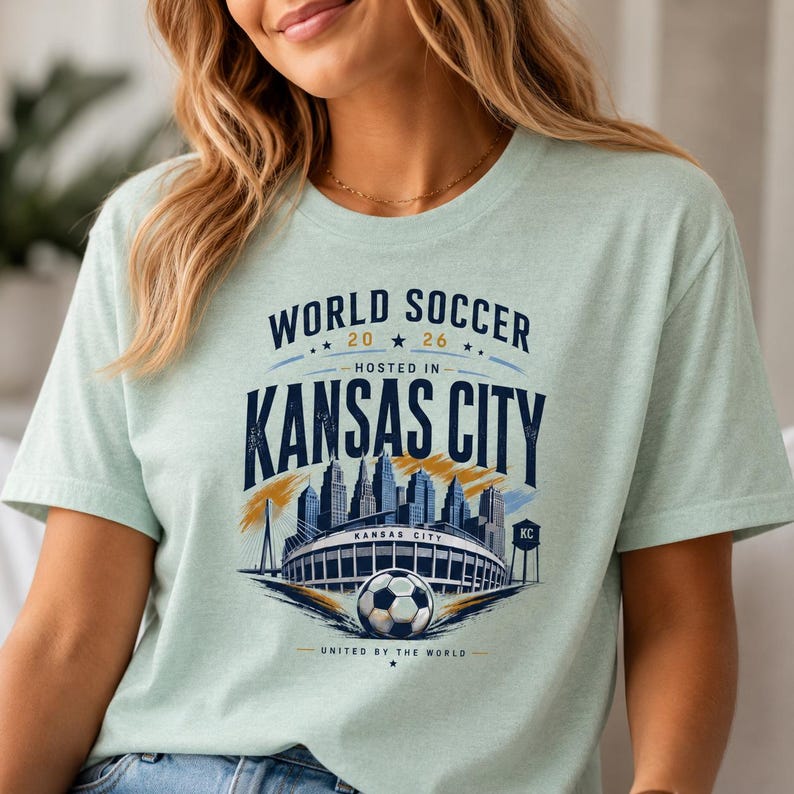 May include: A light green t-shirt featuring a graphic print with the text WORLD SOCCER 2026 HOSTED IN KANSAS CITY UNITED BY THE WORLD. The design includes a stylized city skyline, a stadium, a soccer ball, and a water tower in navy blue and gold.