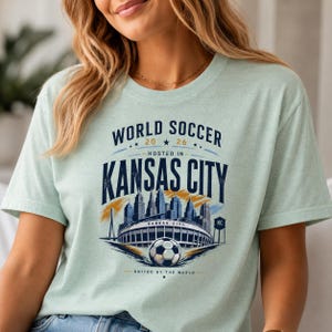 May include: A light green t-shirt featuring a graphic print with the text WORLD SOCCER 2026 HOSTED IN KANSAS CITY UNITED BY THE WORLD. The design includes a stylized city skyline, a stadium, a soccer ball, and a water tower in navy blue and gold.