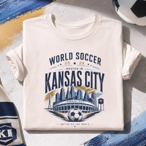 May include: A cream-colored short-sleeve t-shirt featuring a graphic print that reads WORLD SOCCER 2026 HOSTED IN KANSAS CITY UNITED BY THE WORLD. The design includes a stadium, a soccer ball, a city skyline, and a water tower labeled KC.
