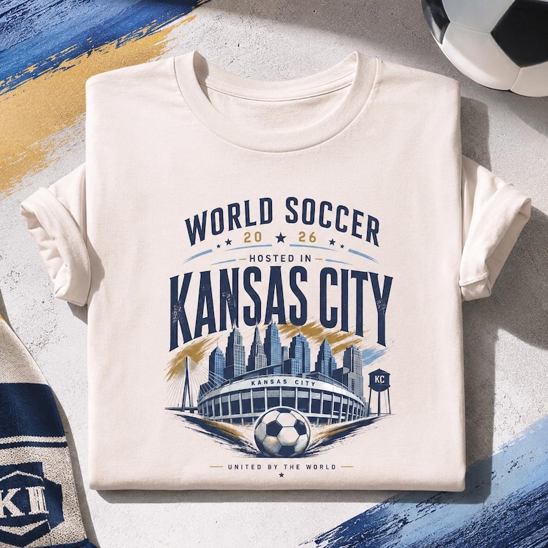 May include: A cream-colored short-sleeve t-shirt featuring a graphic print that reads WORLD SOCCER 2026 HOSTED IN KANSAS CITY UNITED BY THE WORLD. The design includes a stadium, a soccer ball, a city skyline, and a water tower labeled KC.