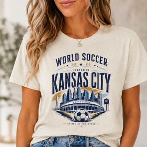 May include: A heathered cream t-shirt featuring a graphic design with the text WORLD SOCCER 2026 HOSTED IN KANSAS CITY. The illustration includes a city skyline, a stadium, a soccer ball, and a water tower with the letters KC printed on it.