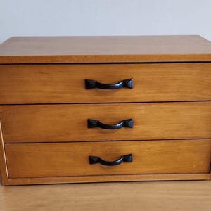 Op de afbeelding: Een houten dressoir met drie lades en zwarte metalen handgrepen. Het dressoir is gemaakt van lichtbruin hout en heeft een eenvoudig ontwerp.