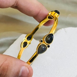 Puede incluir: Un brazalete dorado con piedras grises oscuras en forma de lágrima y piedras negras más pequeñas. El brazalete se sostiene sobre una superficie blanca, con un anillo plateado visible. Joyería para moda o ocasiones especiales.