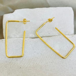 May include: A pair of gold-coloured, rectangular hoop earrings. The earrings have a simple, geometric design with a post closure. The earrings are displayed on a white surface. The earrings are a minimalist style.