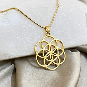 May include: A gold pendant necklace with a geometric floral design. The pendant hangs from a fine gold chain. The necklace is displayed on a white surface and a draped, light-coloured fabric.
