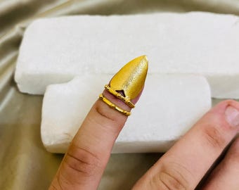 May include: A gold-colored finger armor ring with a pointed tip and two bands. The ring is worn on a finger, with a textured surface and a small triangular cutout near the tip. The jewelry is set against a neutral background.
