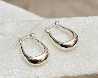 May include: A pair of polished silver hoop earrings. The earrings have a smooth, rounded design and a secure clasp closure. The earrings are displayed on a white, textured surface.