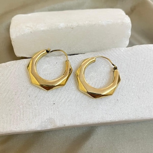 Op de afbeelding: Een paar goudkleurige oorringen met een geometrisch, facetgeslepen ontwerp. De oorbellen hebben een gepolijste afwerking en een scharniersluiting. De oorbellen worden gepresenteerd op een wit, getextureerd oppervlak.