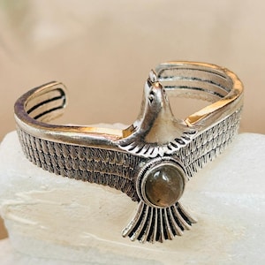 Könnte beinhalten: Silberfarbenes Armreif-Armband mit einem Adler-Design. Das Armband hat detaillierte Feder- und Flügelmuster mit einem runden, hellfarbigen Stein in der Mitte. Der Kopf des Adlers ist deutlich sichtbar und verleiht dem Schmuck ein kühnes Aussehen.