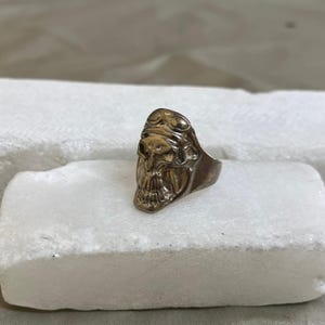 以下が含まれることがあります： 精巧なディテールが施されたブロンズ色のスカルリング。リングは、頭飾りをつけたスカルのデザインが特徴です。リングは白い表面に置かれています。ユニークなジュエリーです。
