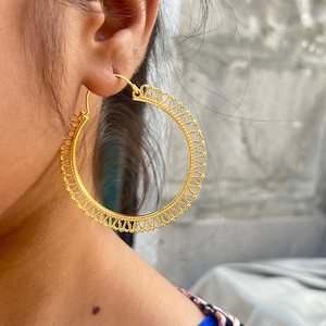 May include: Gold-colored hoop earrings with an intricate, openwork design. The earrings have a circular shape with a detailed pattern of small, connected teardrop shapes around the outer edge. The earrings are shown on a person's ear.