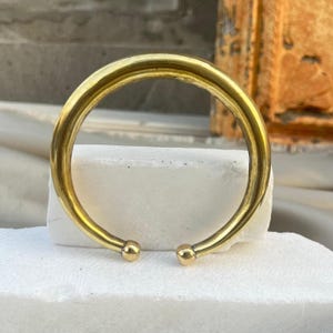 Peut inclure: Un bracelet manchette ouvert de couleur or avec un design arrondi. Le bracelet a une finition lisse et polie et de petits accents sphériques aux extrémités ouvertes. Le bijou est présenté sur une surface blanche.