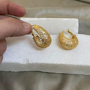 Peut inclure: Deux boucles d'oreilles créoles dorées avec un motif ajouré. Les boucles d'oreilles présentent des motifs complexes. L'une est tenue par une main, l'autre repose sur une surface blanche. Les boucles d'oreilles sont en métal brillant.