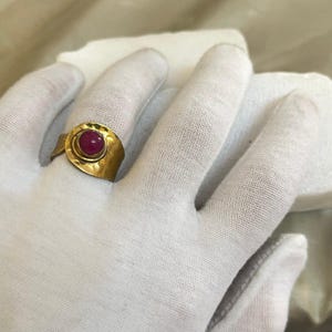 Op de afbeelding: Een goudkleurige ring met een grote, ronde, rode edelsteen. De ring heeft een getextureerde band en een cirkelvormige zetting rond de edelsteen. De ring zit op een gehandschoende hand.