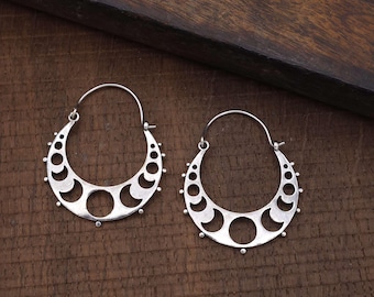 May include: A pair of silver-colored hoop earrings. Each earring features a crescent moon design with cut-out shapes and small, round embellishments. The earrings are displayed on a dark brown wooden surface.