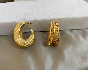 May include: A pair of gold-coloured hoop earrings. One earring has a textured, dotted surface, the other features a geometric design. The earrings are presented on a light grey surface, suggesting a jewellery or fashion theme.