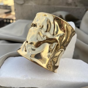 Può includere: Un bracciale a polsino largo color oro con un aspetto martellato e strutturato. Il bracciale ha una forma leggermente irregolare e riflette la luce, creando riflessi e ombre. Il gioiello è esposto su una superficie bianca.