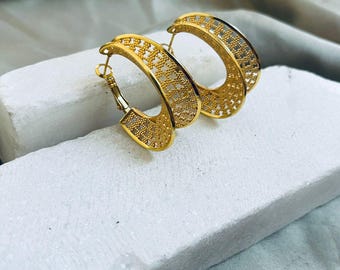 May include: A pair of gold-colored hoop earrings. The earrings have a detailed, openwork design with a repeating pattern. The hoops are displayed on a white surface against a neutral background.