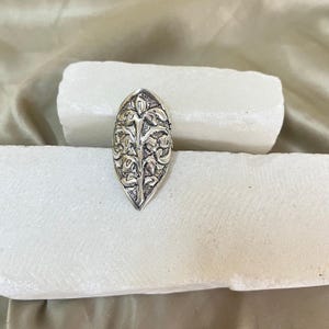 Puede incluir: Un anillo plateado con forma alargada, similar a una hoja. El anillo presenta un intrincado diseño floral en relieve en toda su superficie. El anillo se muestra sobre una superficie blanca y rectangular, sobre un fondo neutro.