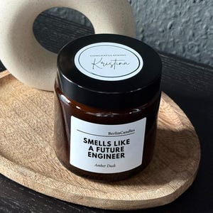 Könnte beinhalten: Eine Kerze in einem braunen Glas mit schwarzem Deckel auf einem Holztablett. Auf dem Etikett steht "Smells Like A Future Engineer". Eine cremefarbene Vase mit kreisförmiger Öffnung enthält Trockenblumen. Die Kerze duftet nach "Amber Dusk".