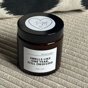 Könnte beinhalten: Ein dunkelbraunes Glas-Kerzenglas mit schwarzem Deckel. Das Etikett lautet "Smells like one year, still obsessed" und "Amber Dusk". Die Kerze steht auf einer neutralen Oberfläche, mit einem schwarz-weiß gemusterten Kissen im Hintergrund.